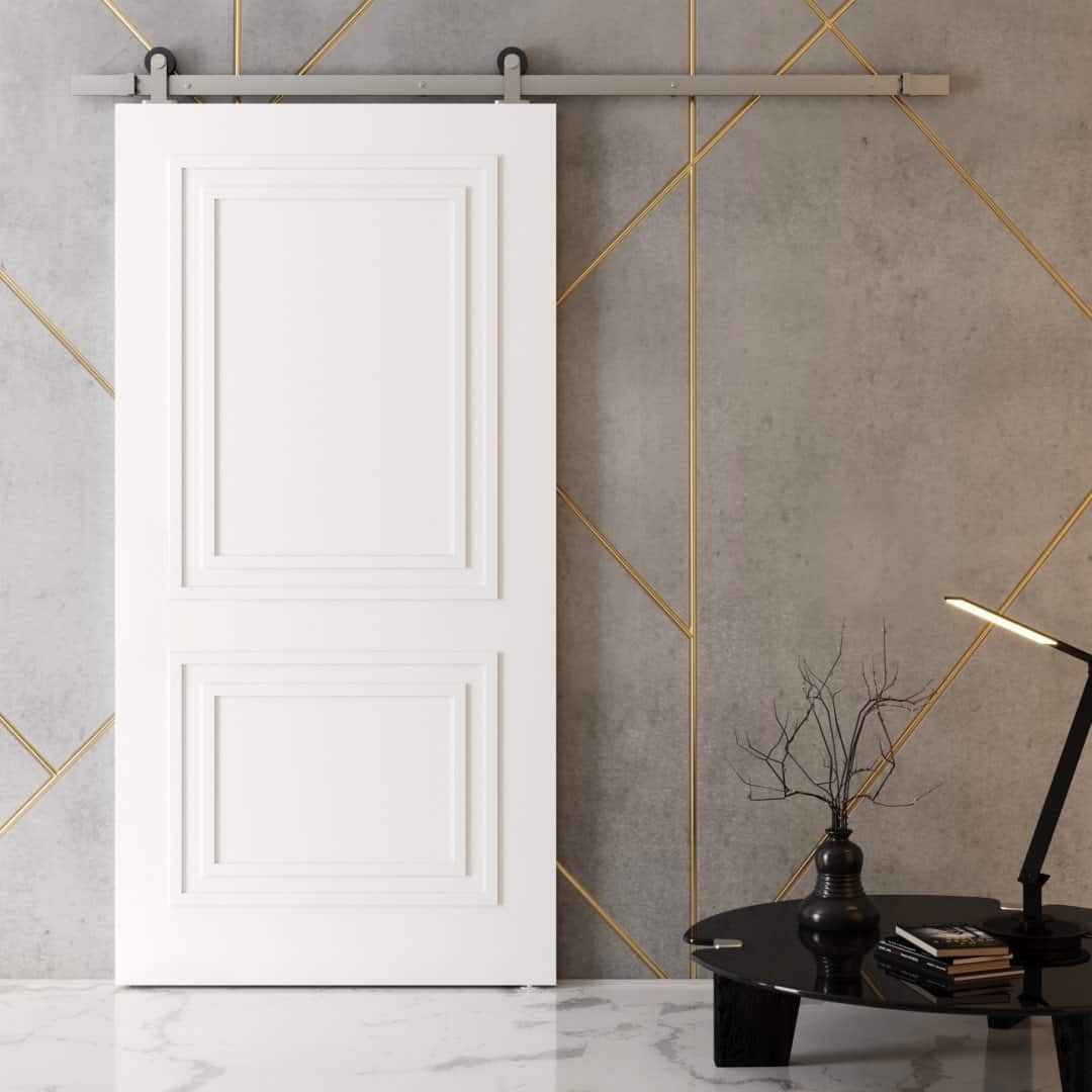 An Atelier Barn Door in modern white with gold accents, installed on a concrete wall, is accompanied by a black side table adorned with a lamp, tree branch decoration, and stacked books.