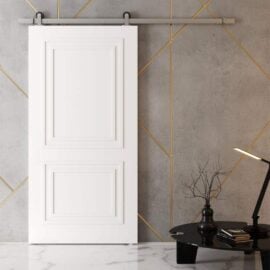 An Atelier Barn Door in modern white with gold accents, installed on a concrete wall, is accompanied by a black side table adorned with a lamp, tree branch decoration, and stacked books.