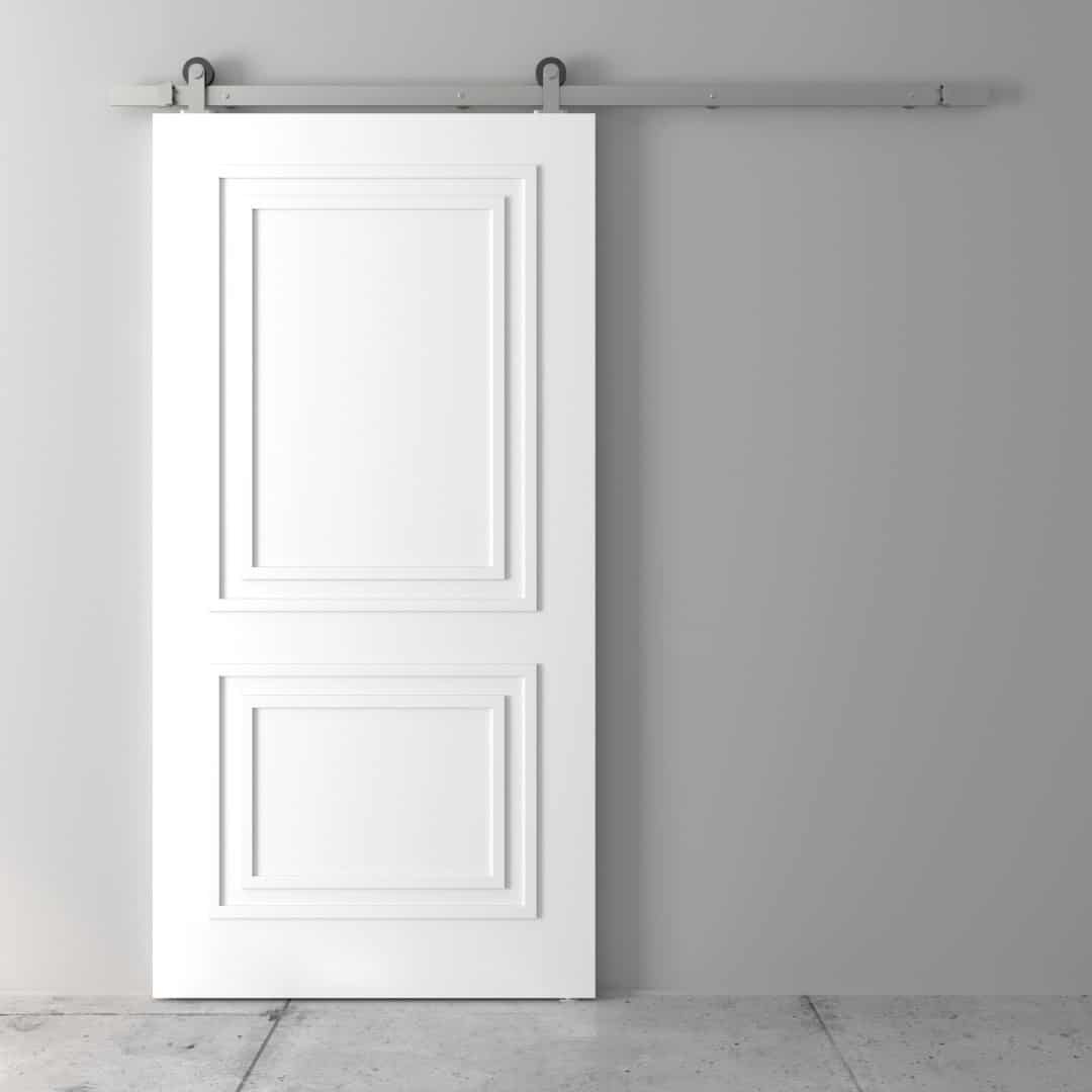 The Atelier Barn Door, featuring raised rectangular panel designs, is mounted on a metal track against a grey wall.