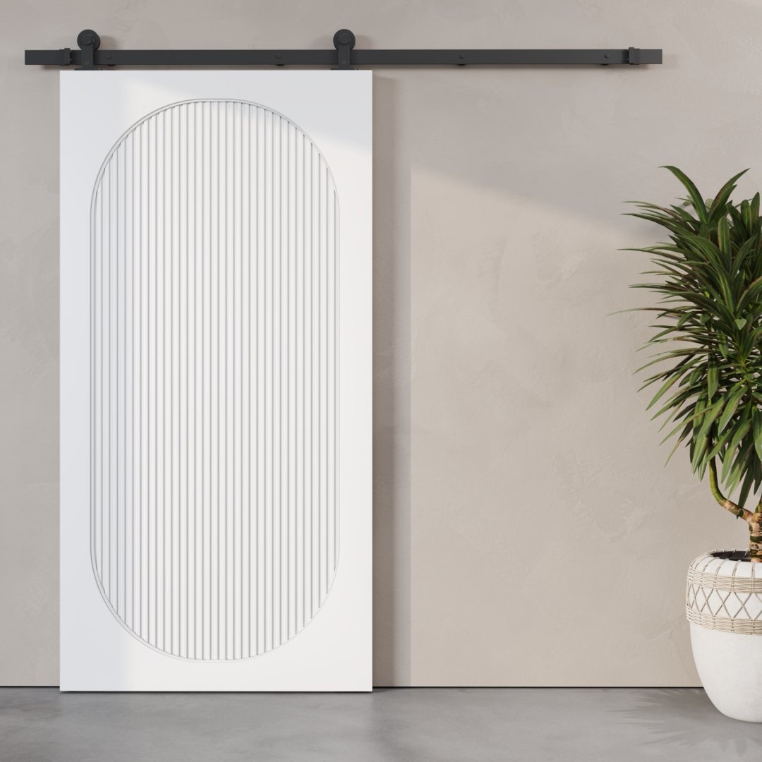 The Celine Oval Fluted Panel Barn Door, featuring a white finish and an oval design, is mounted on a black rail against a beige wall. A potted plant is placed to the right of the door.