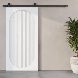 The Celine Oval Fluted Panel Barn Door, featuring a white finish and an oval design, is mounted on a black rail against a beige wall. A potted plant is placed to the right of the door.