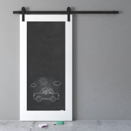 The Chalkboard Barn Door, with its white frame, showcases a hand-drawn image of a car with smiling faces, a sun, and clouds. Chalk and an eraser rest on the floor beneath the door.