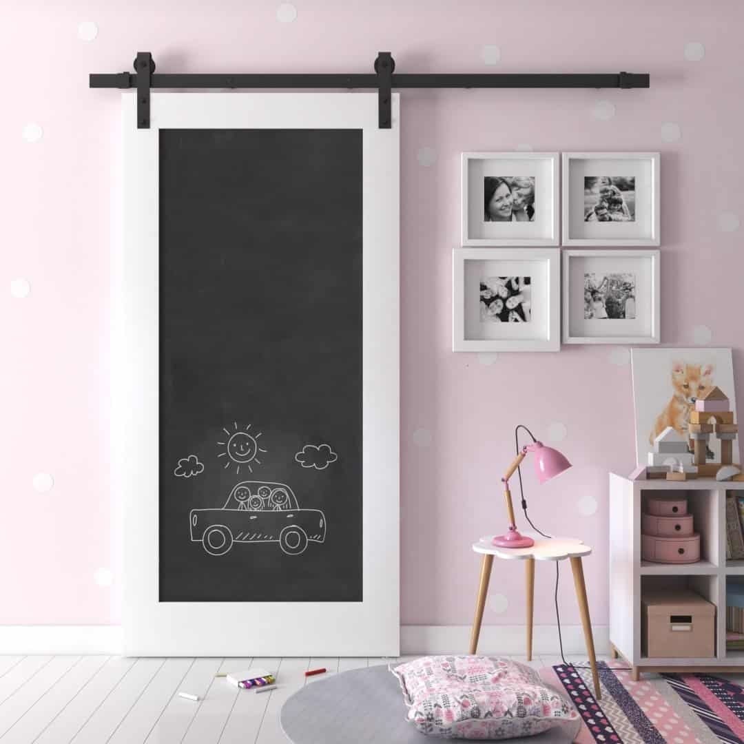A children's room featuring a pink polka-dot wall, the Chalkboard Barn Door with a drawing of a car, framed pictures, a small bookshelf, a pink lamp, and a cushion on the floor.
