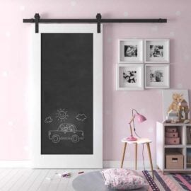 A children's room featuring a pink polka-dot wall, the Chalkboard Barn Door with a drawing of a car, framed pictures, a small bookshelf, a pink lamp, and a cushion on the floor.
