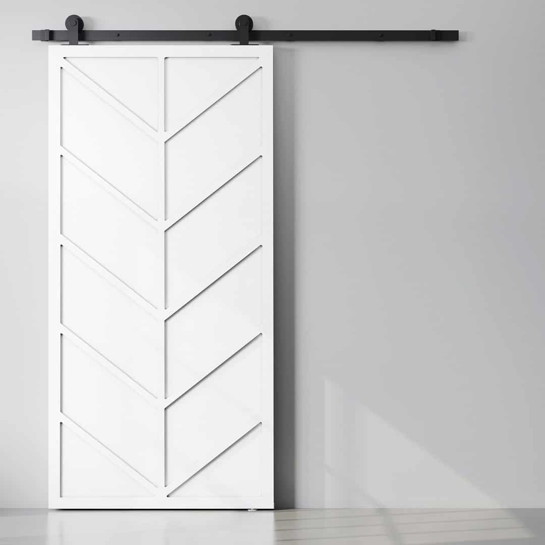 Canal-Barn-Door-White-1.jpg The Canal Barn Door, featuring a chevron pattern and mounted on a black track, is set against a grey wall.