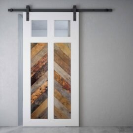 The Lake House Barn Door, featuring a white finish with two small glass panels at the top and a chevron-patterned wooden design in the middle, stands against a light gray wall.