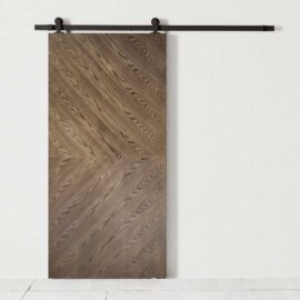 The Bianco Barn Door, featuring a chevron pattern on its wooden surface, is mounted on a black metal rail against a white wall.