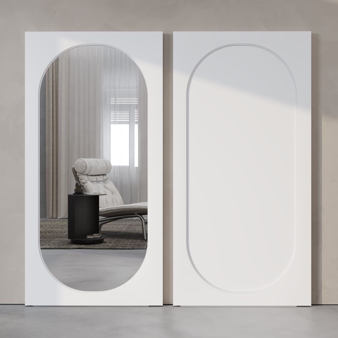 A modern interior features two large, vertical rectangular frames on a wall, one with the Celine Oval Mirror Barn Door reflecting a lounge chair and side table, and the other with a plain, decorative design.