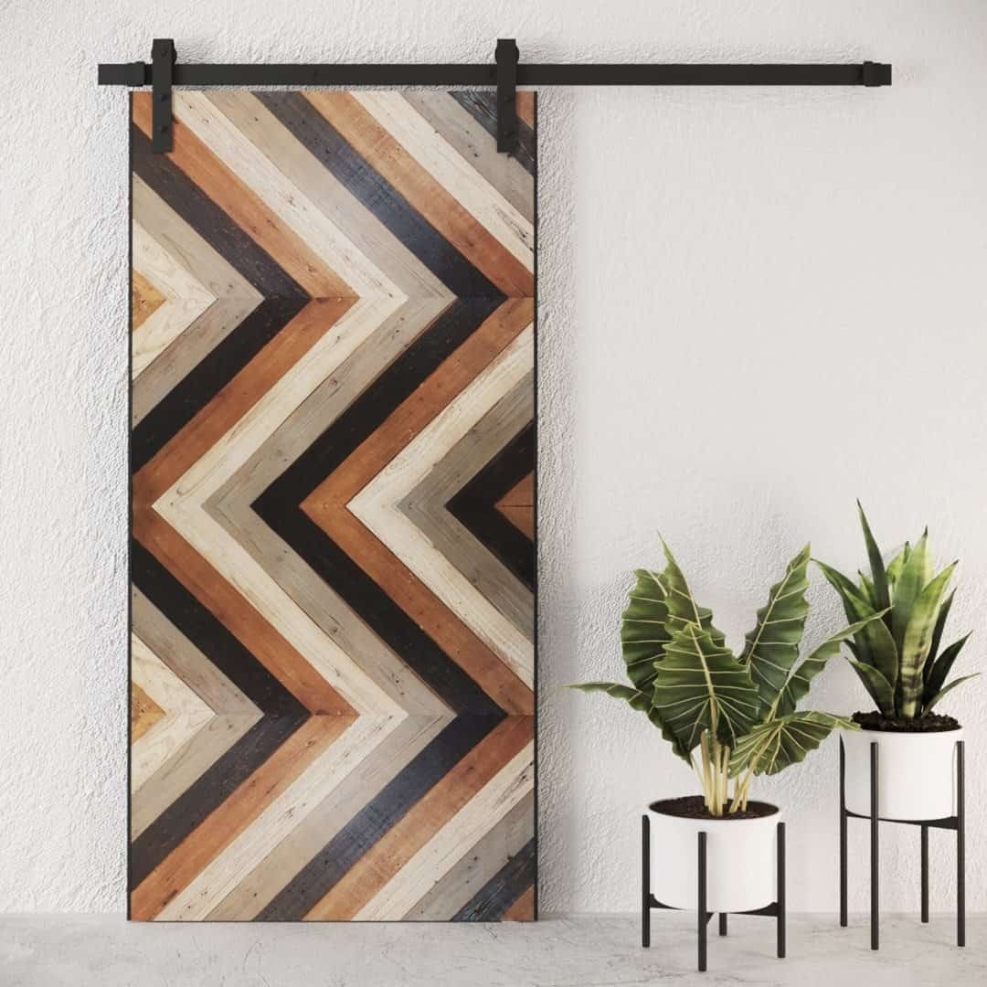 A Cali Chevron Barn Door featuring a chevron pattern in neutral tones, positioned beside two potted indoor plants.
