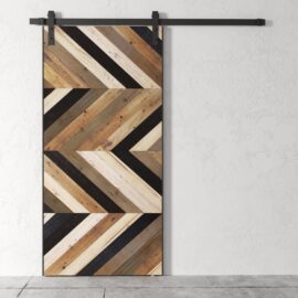The Bungalow Chevron Barn Door, showcasing a chevron pattern made from various shades of wood, is mounted on a white wall with a black metal track.