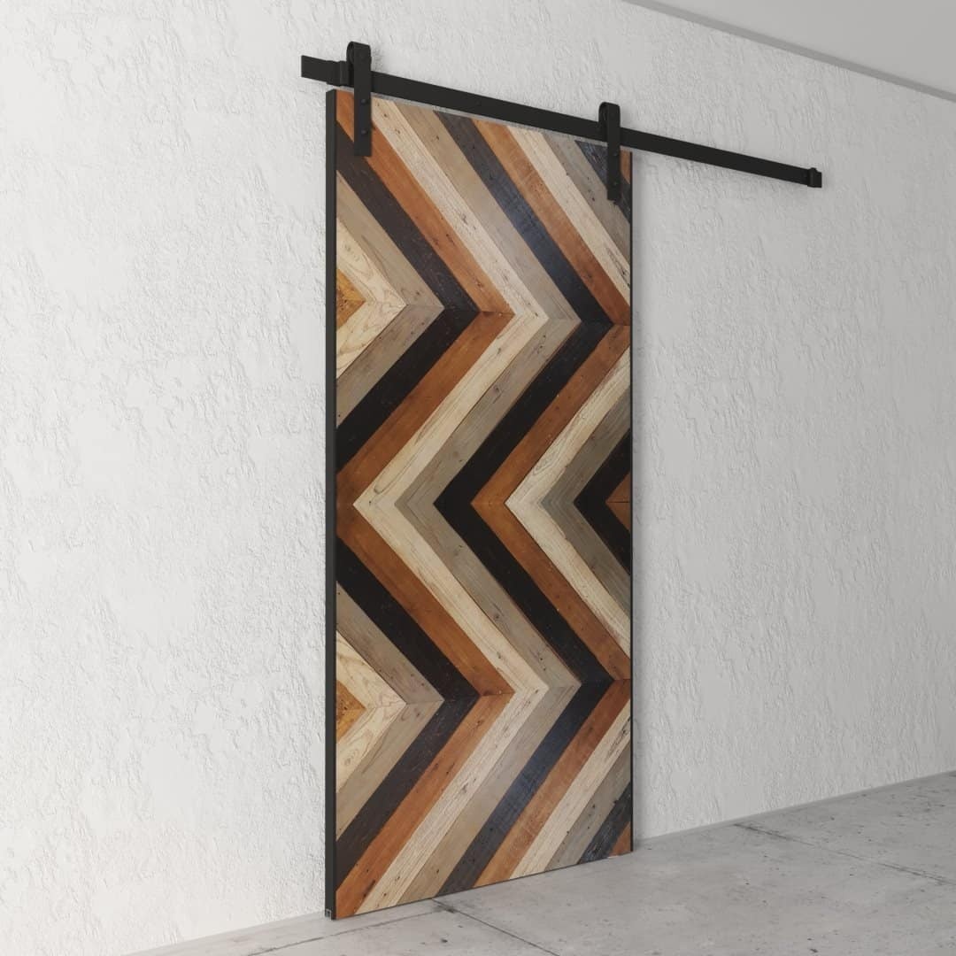 Urban-Woodcraft-Chevron-Barn-Door-Cali-3.jpg The Cali Chevron Barn Door, featuring a striking chevron wood pattern in various shades of brown, is beautifully mounted on a sleek black rail against a white textured wall.