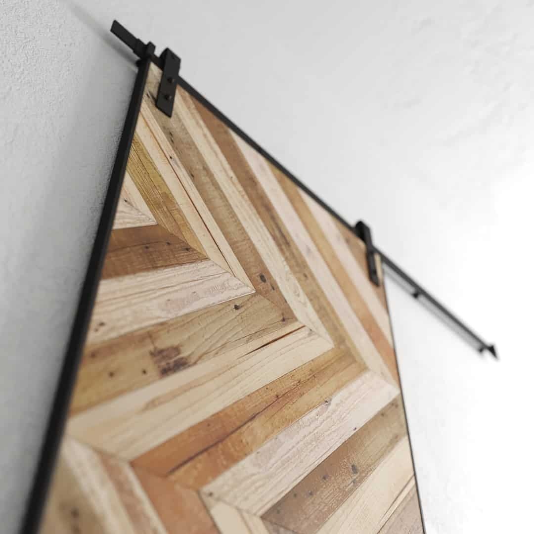 The Oasis Chevron Barn Door, crafted from light and dark wood panels in a chevron pattern, is mounted on a black metal rail against a white wall.