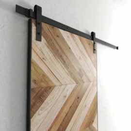 The Scandi Chevron Barn Door, featuring a wooden chevron pattern, is mounted on a black metal track against a white wall.