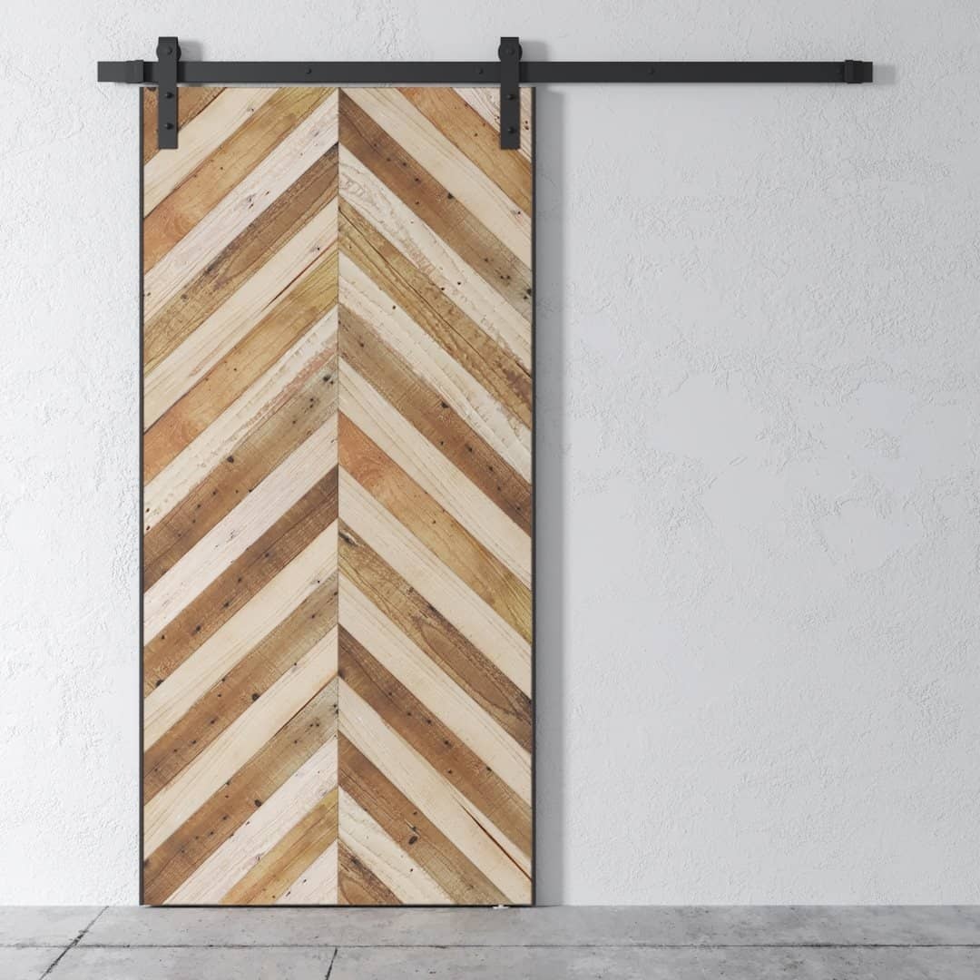 The Bohemian Herringbone Barn Door, featuring a distinctive wooden herringbone pattern, is mounted on a dark metal rail against a white textured wall. The floor beneath appears to be concrete.