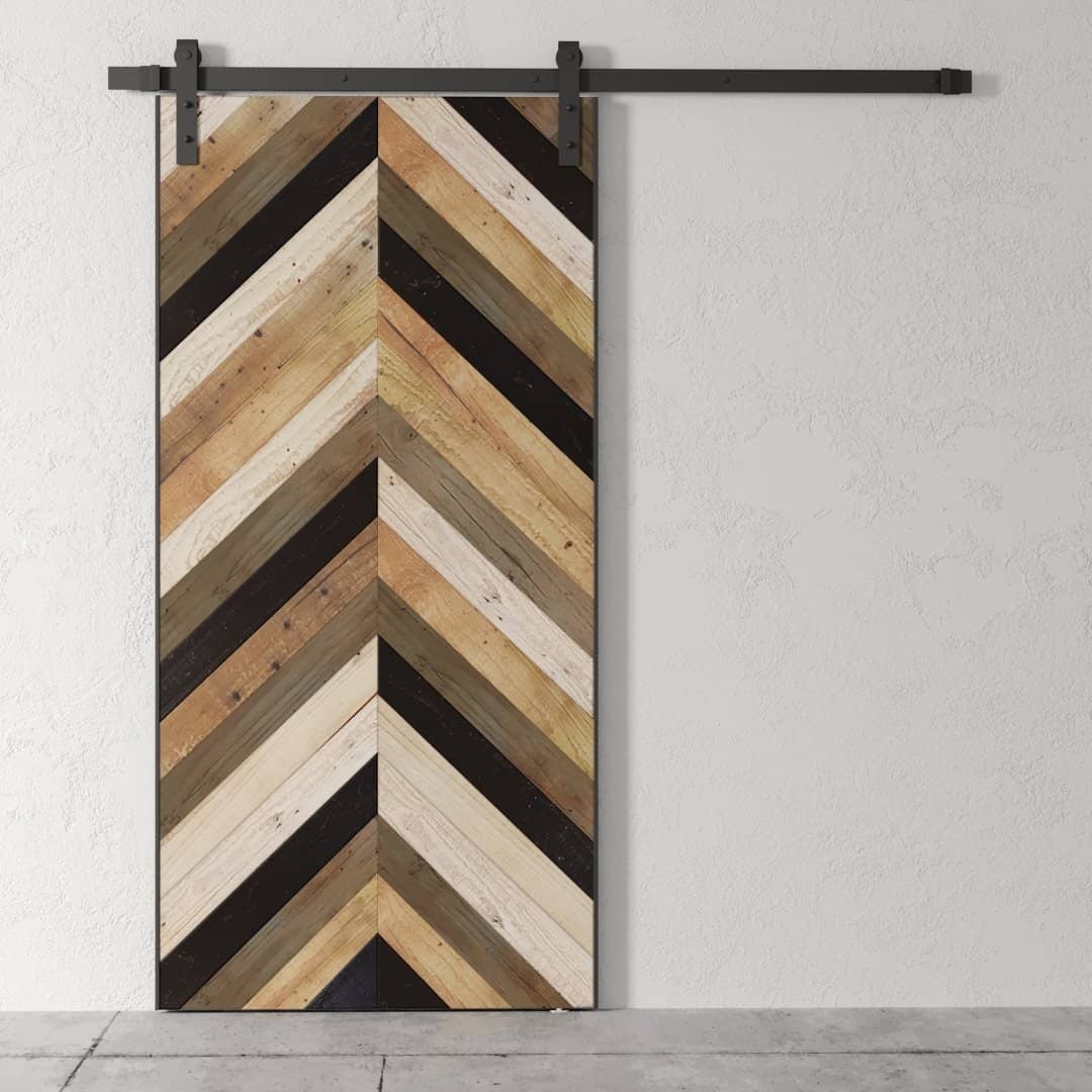 A Wilderness Herringbone Barn Door, featuring a chevron pattern crafted from alternating dark and light wood slats, mounted on a metal rail, against a plain white wall.