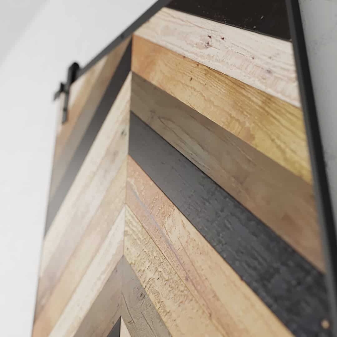Close-up of the Wilderness Herringbone Barn Door with its distinctive wooden chevron-patterned design featuring alternating colors and textures in diagonal strips. The pattern showcases an array of light, dark, and natural wood tones.