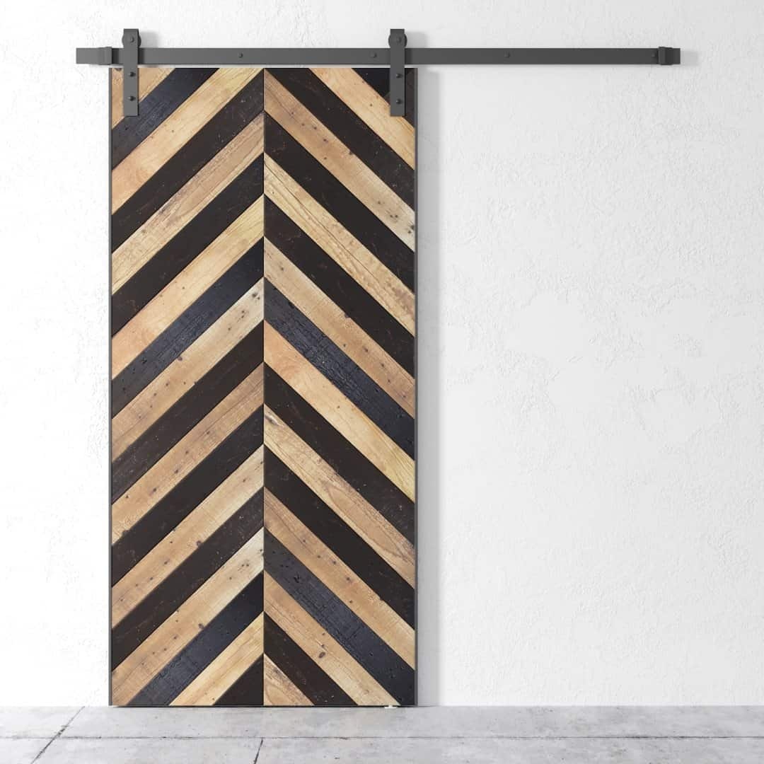 The Alchemist Herringbone Barn Door, designed with alternating light and dark planks in a herringbone pattern, stands out against a plain white wall.
