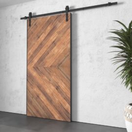 The Lucas Reclaimed Wood Arrow Barn Door, featuring a distinctive herringbone pattern, is mounted on a black metal rail against a white wall. A green plant is partially visible on the right side.