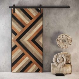 The Cali Malibu Barn Door, featuring a chevron pattern, stands beside two ornate stone sculptures on a stand against a textured gray wall.