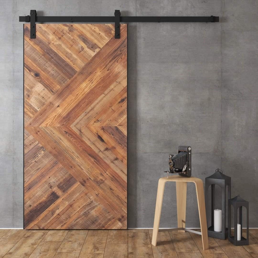 The Malibu Barn Door, a rustic wooden sliding barn door, is mounted on a gray wall. Nearby, a wooden stool holds a vintage camera, and two lanterns with white candles sit on the floor.