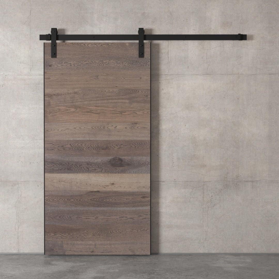 Urban-Woodcraft-Oak-Panel-Barn-Door-Grey-1.jpg An Oak Panel Barn Door mounted on a concrete wall with black metal hardware.