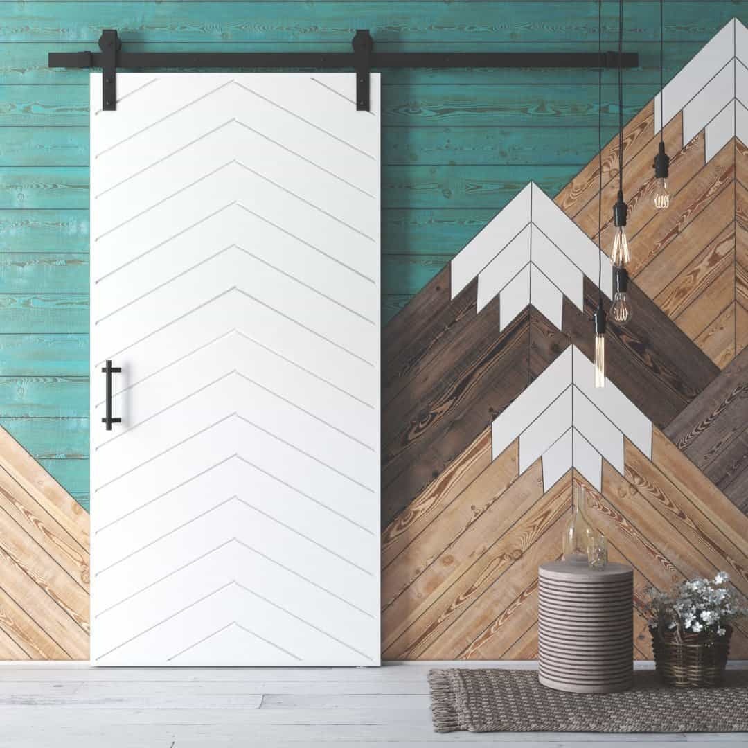 The Osaka Barn Door, featuring a chevron design, slides on a black rail against a teal and wood patterned accent wall, complemented by hanging light bulbs and a small cylindrical woven stool.