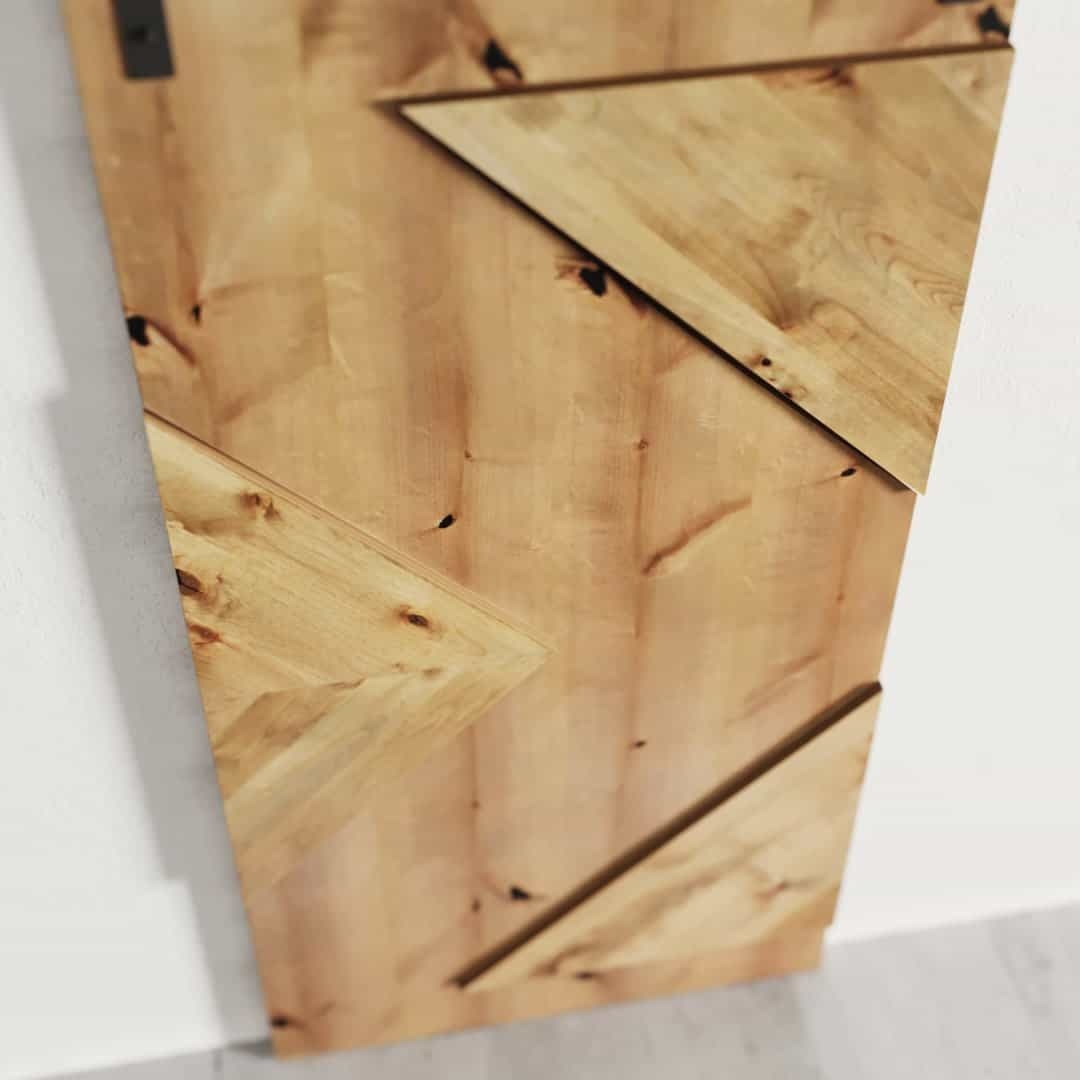 Urban-Woodcraft-Raised-Arrowhead-Barn-Door-Knotty-Alder-3.jpg Introducing the Raised Arrow Head Barn Door: a stunning wooden door featuring a geometric design highlighted by triangular wooden panels. This light brown door showcases a natural wood grain pattern, making it an elegant addition to any space.