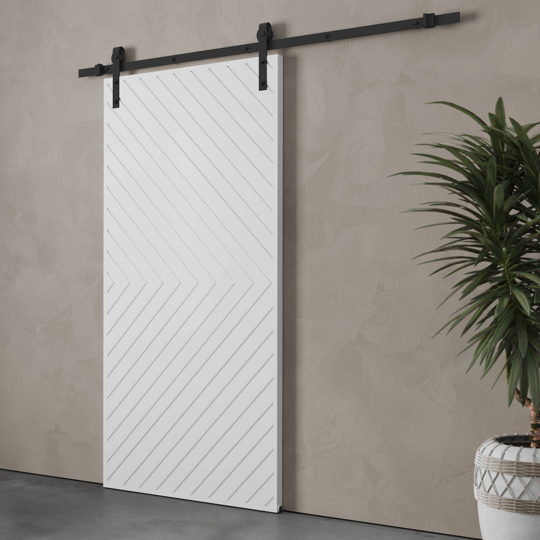 The Venice Arrow Barn Door, featuring a white finish with diagonal line patterns, is mounted on a sleek black rail against a beige textured wall. A potted plant stands nearby.