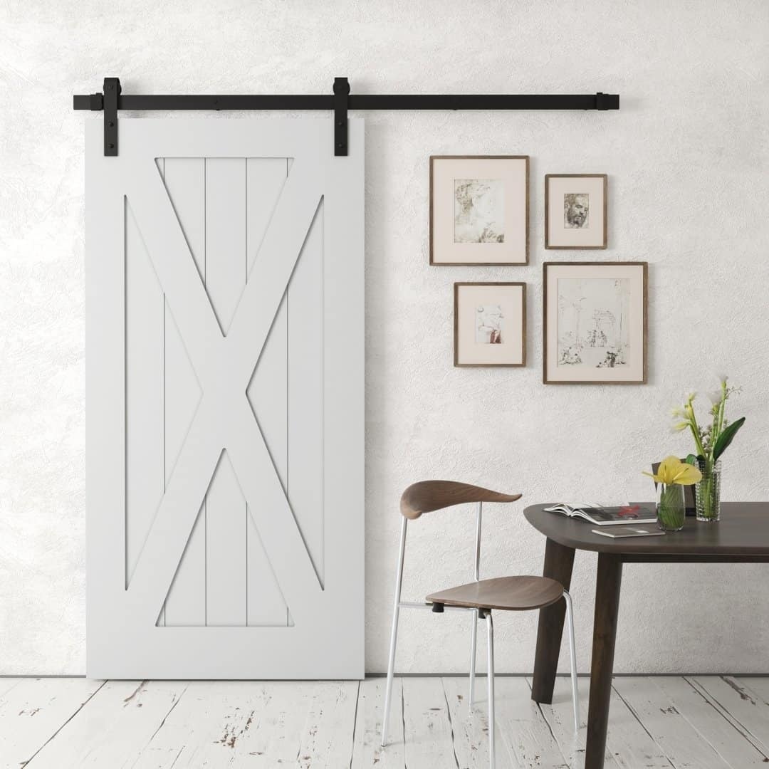 An X-Panel Barn Door in white is mounted on a black rail. Nearby, there's a table with a vase of yellow flowers and two brown chairs. Three framed pictures hang on the wall.