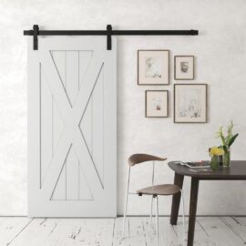 An X-Panel Barn Door in white is mounted on a black rail. Nearby, there's a table with a vase of yellow flowers and two brown chairs. Three framed pictures hang on the wall.
